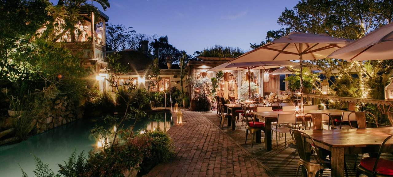 A lantern-lit lodge terrace and pool at dusk, tables set under umbrellas